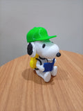 Figura do Snoopy sentado com boné verde, mochila amarela e livro azul